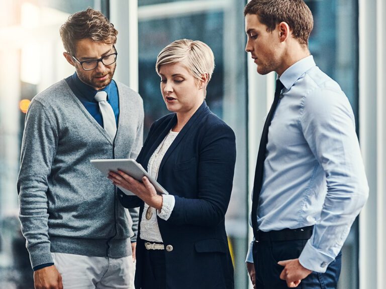 Three people are standing and looking at a tablet that one person is holding. They are dressed in business attire and appear to be having a discussion.