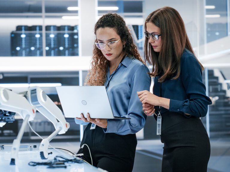 Two women in a modern office setting look at a laptop screen together, surrounded by technology and equipment. Both wear glasses and business attire.