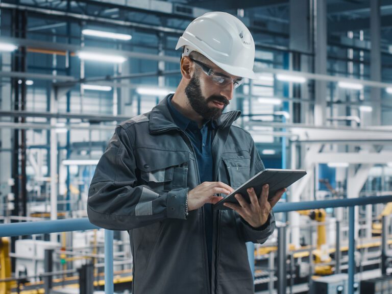 A man in a hard hat and safety glasses uses a tablet in an industrial setting.