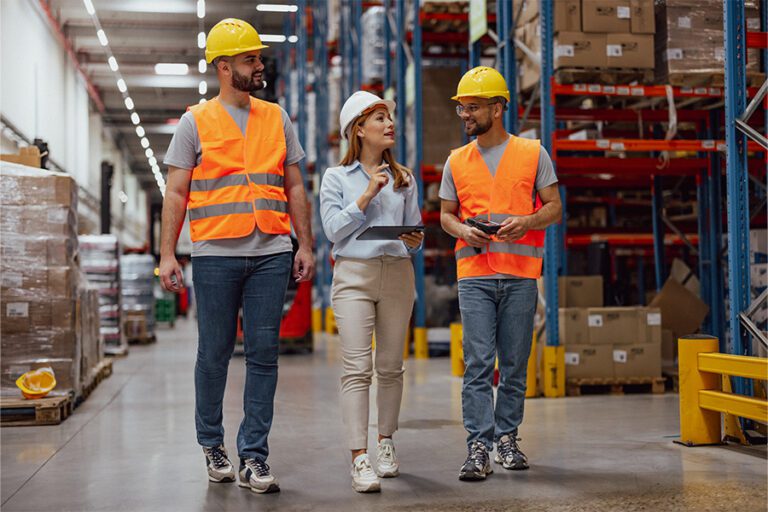 Three warehouse workers wearing safety vests and helmets walk and discuss while holding a clipboard and a tablet in a large warehouse.