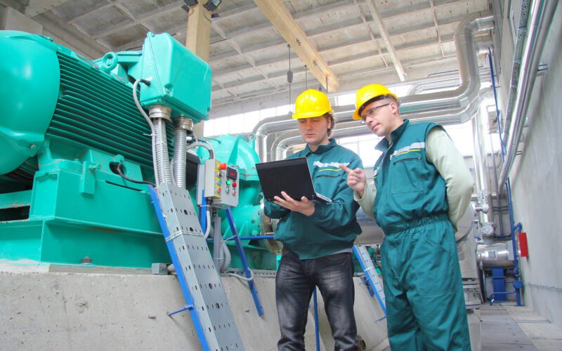 Two workers wearing hard hats and green uniforms inspect industrial machinery, as one holds a laptop, inside a factory with exposed pipes and machinery in the background.