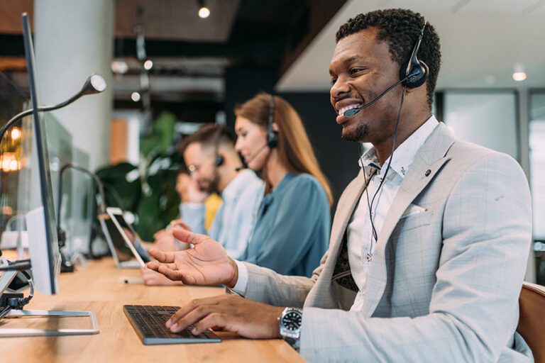 Three customer service representatives wearing headsets, including one man in the foreground and two people in the background, are working at computers in a modern office setting.