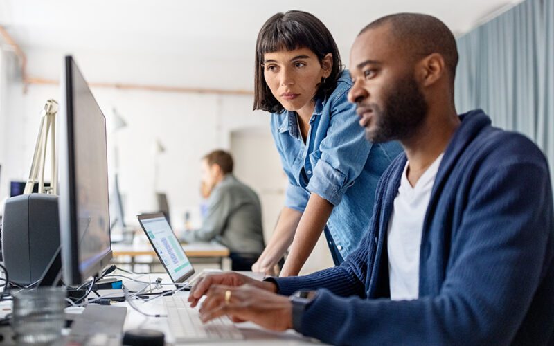 Two individuals work together at a desk with laptops and monitors in a modern office setting. One person stands and leans over while the other sits and types. Another individual works in the background.