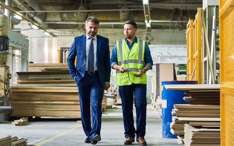 Two men walking through a warehouse; one is in a blue suit and the other in a reflective vest, conversing. Shelves and industrial equipment are visible in the background.