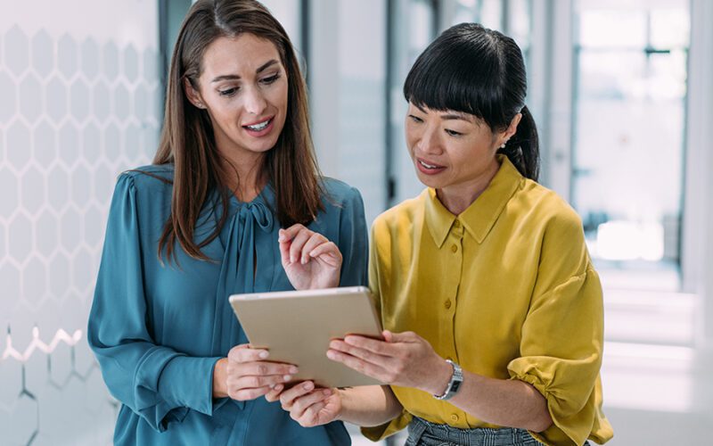 Two women are standing in a modern office hallway, looking at a tablet. One woman wears a blue blouse and the other wears a yellow blouse. They are engaged in discussion.