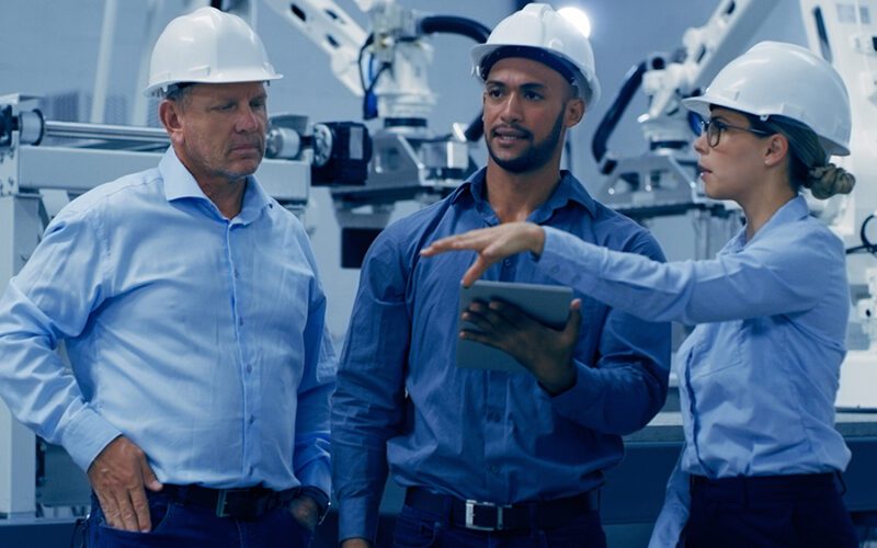 Three individuals wearing hard hats and safety glasses discuss something on a tablet in an industrial setting with machinery in the background.