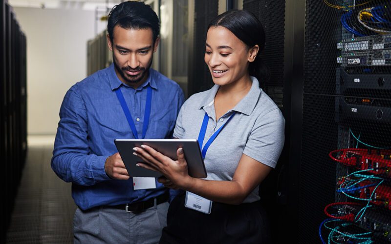 Two people in a server room, each wearing a badge, review information on a tablet. One person is holding the tablet while the other looks on. Server racks are visible in the background.