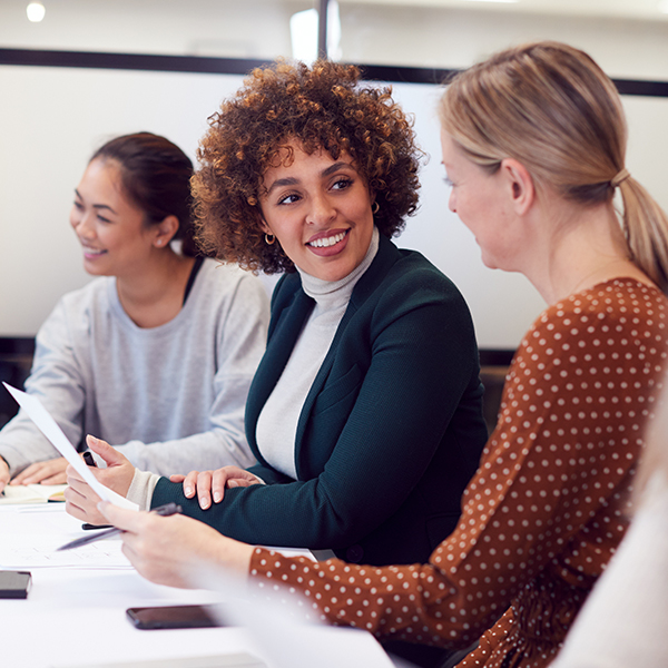 Three women are sitting at a table in an office, holding papers and smiling at each other.