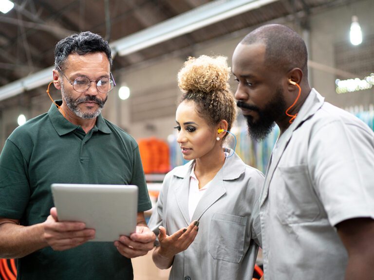 Three factory workers wearing safety gear discuss something while looking at a tablet.