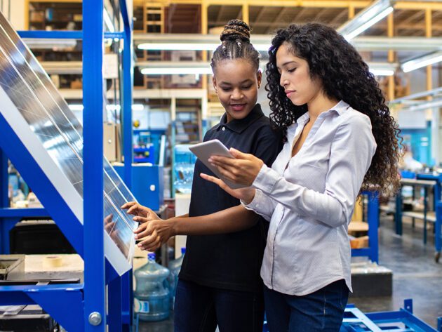 Two women in a factory setting, one using a touchscreen on machinery, and the other holding a tablet.