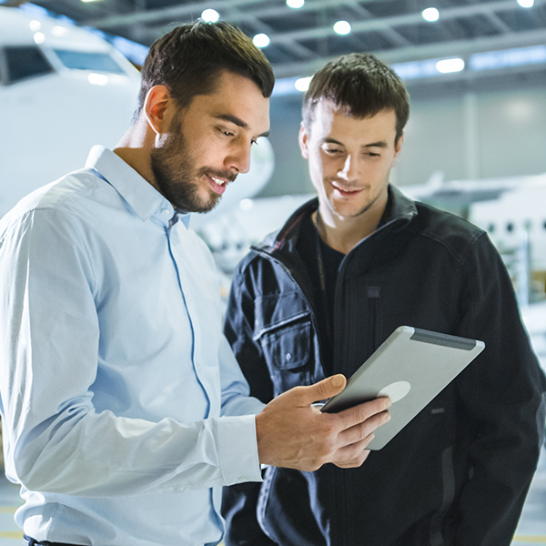 Two men stand in front of an airplane, looking at a tablet.