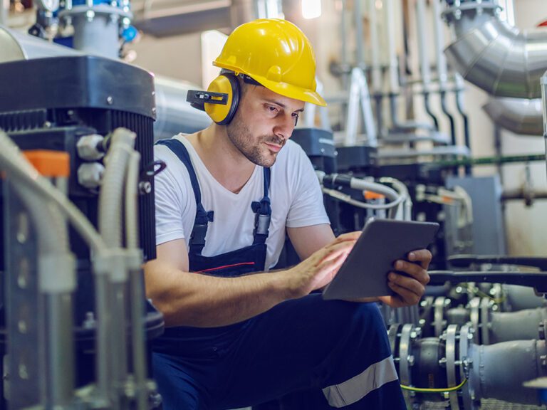 A worker in a hard hat and earmuffs uses a tablet in an industrial setting with pipes and machinery.