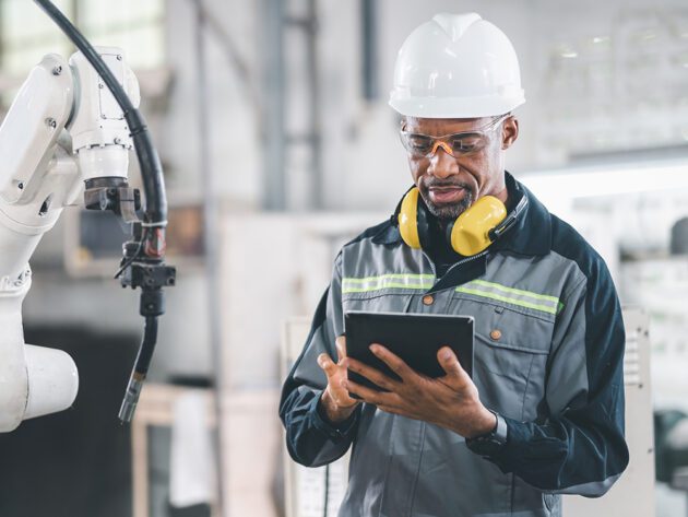 A man in safety gear examines a tablet near industrial machinery in a factory setting.