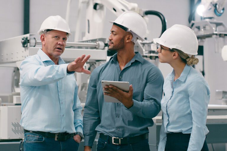 Three people in hard hats and blue shirts stand in a factory setting, discussing with one holding a tablet. Machinery is visible in the background.