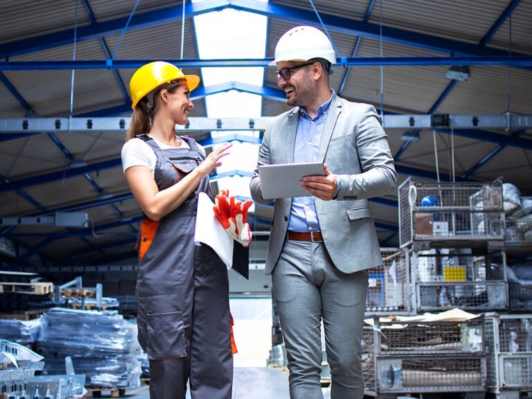 A worker in a yellow hard hat and a person in a suit with a white hard hat converse in a warehouse. The person in the suit holds a tablet.