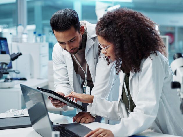 Two scientists in lab coats discuss information on a tablet and a laptop in a laboratory setting.