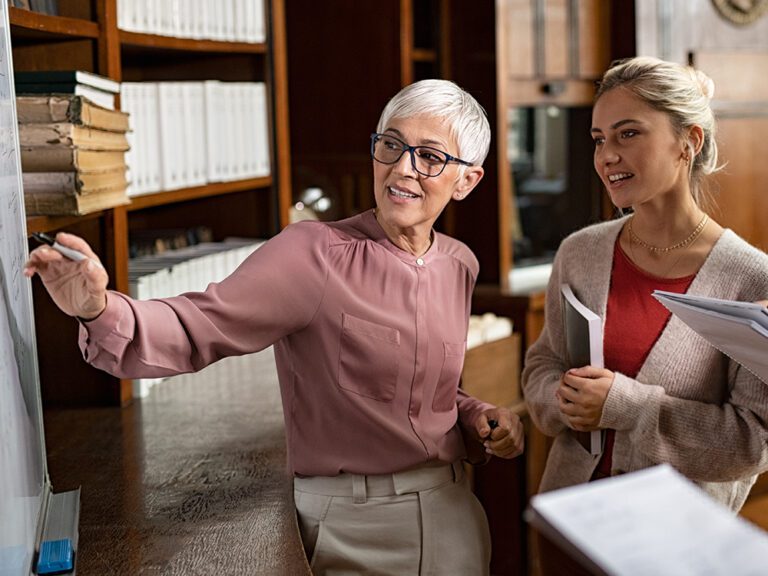 An older woman explains a concept on a whiteboard to a younger woman in an office setting.