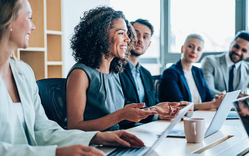 Five people sit at a conference table with laptops. A woman in the center speaks, and the others listen attentively.