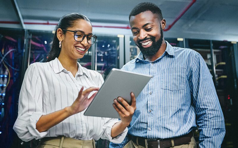 Two people in a server room smile while looking at a tablet. The woman holds the device and wears glasses; both are dressed in business casual attire.
