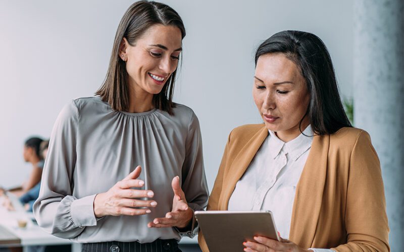 Two women in business attire discuss something while looking at a tablet. One woman gestures with her hands, and the other focuses on the device.