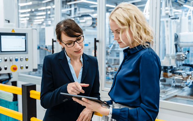 Two women in a factory setting discuss information displayed on a tablet. One woman is pointing at the screen.