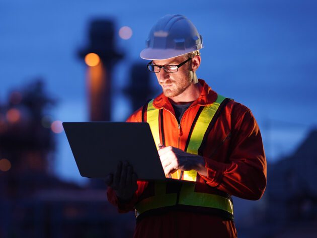 A worker in a hard hat and safety vest uses a laptop outdoors at dusk, with industrial structures blurred in the background.