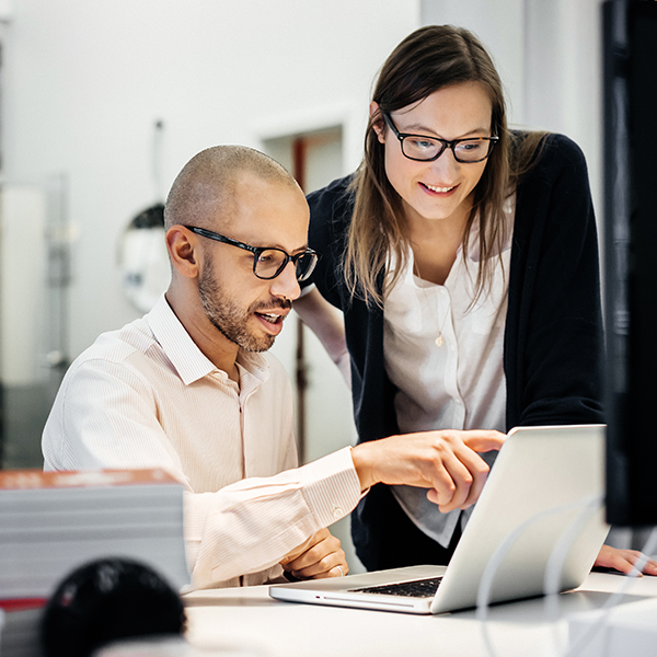 Two people, one seated and one standing, collaborate while looking at a laptop in an office setting. The seated person is pointing at the laptop screen.