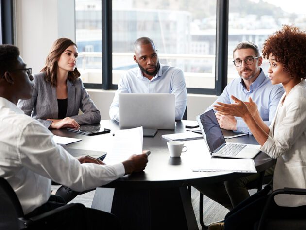 Five people in a conference room sit around a table with laptops and documents, engaged in a discussion.