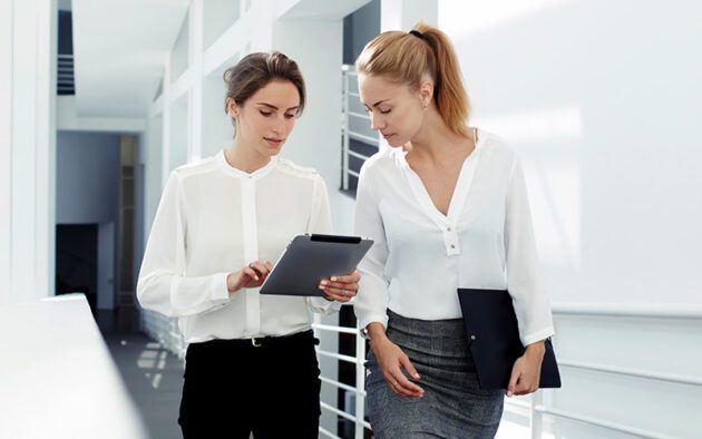 Two women in an office corridor discuss content on a tablet. Both wear white shirts, and one holds a folder.