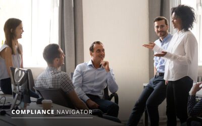 Five people are engaged in a discussion in an office setting. One person is standing and speaking, while the others are sitting, listening attentively. A "Compliance Navigator" logo is visible.