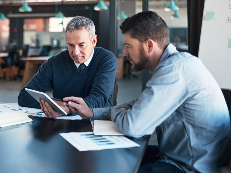 Two men sit at a table reviewing information on a tablet, with documents and charts spread out in front of them. They are in a modern office setting.