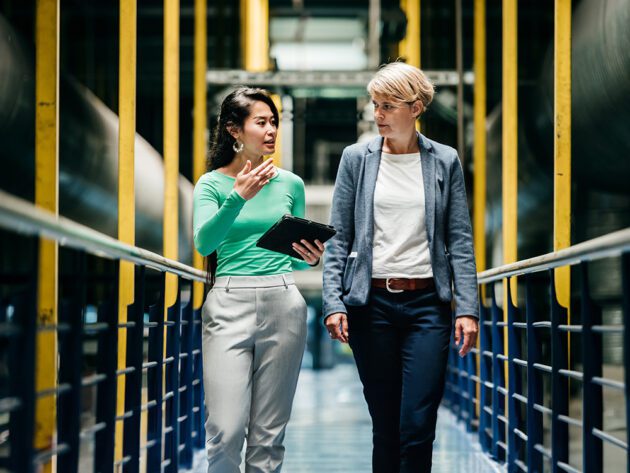 Two people walking and discussing something in an industrial setting. One holds a tablet.