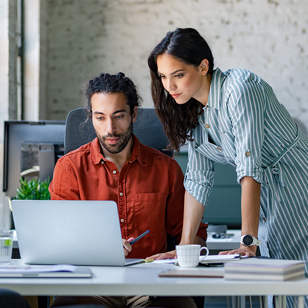 Two people working at a desk with a laptop, papers, and coffee cups in an office setting.