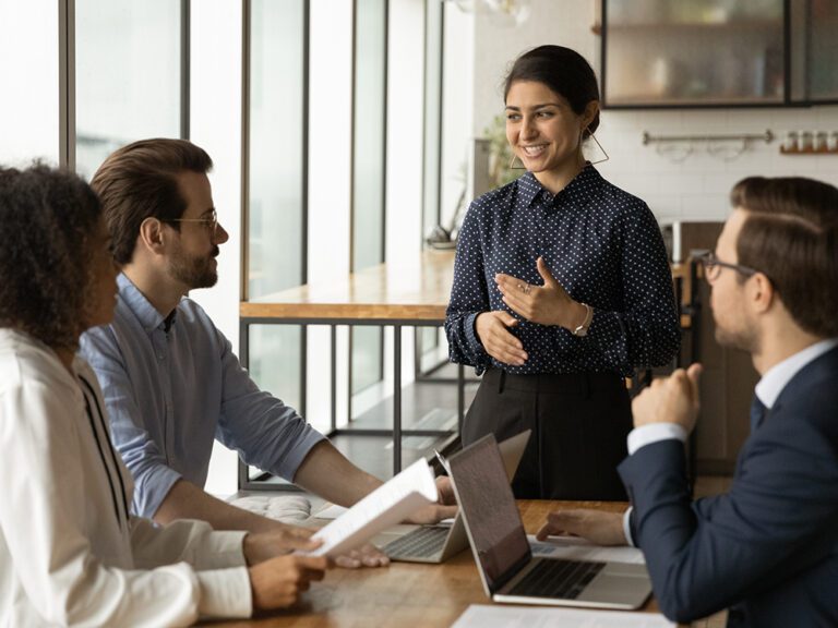 A woman stands and speaks to three seated colleagues at a table with laptops and documents in a modern office.