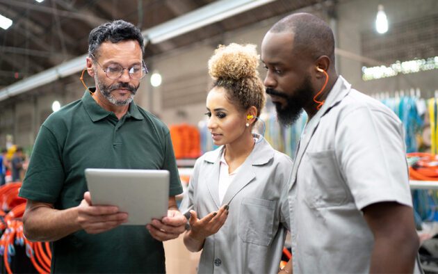 Three people are discussing while looking at a tablet in an industrial setting. They all wear ear protection.