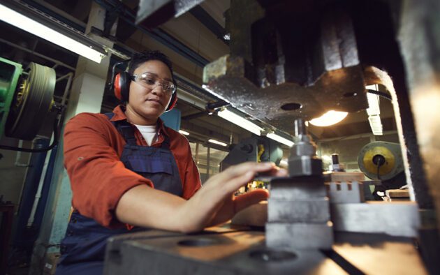 Person operating a large industrial machine, wearing safety goggles and earmuffs, in a workshop setting.