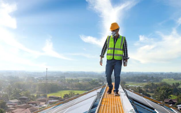 Person in a safety vest and hard hat walking on a roof under a clear blue sky with clouds.