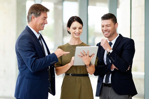 Three people stand indoors, smiling and looking at a tablet. Two men in suits are on either side of a woman in a green dress.