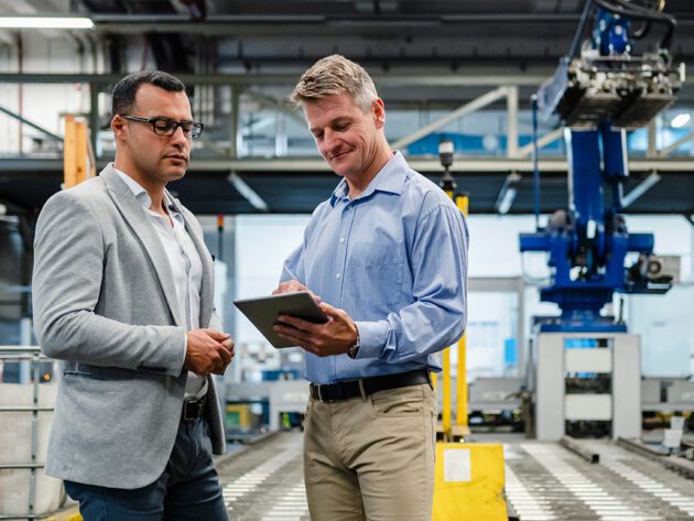 Two men in a factory setting, with one holding a tablet. Machinery and equipment are visible in the background.