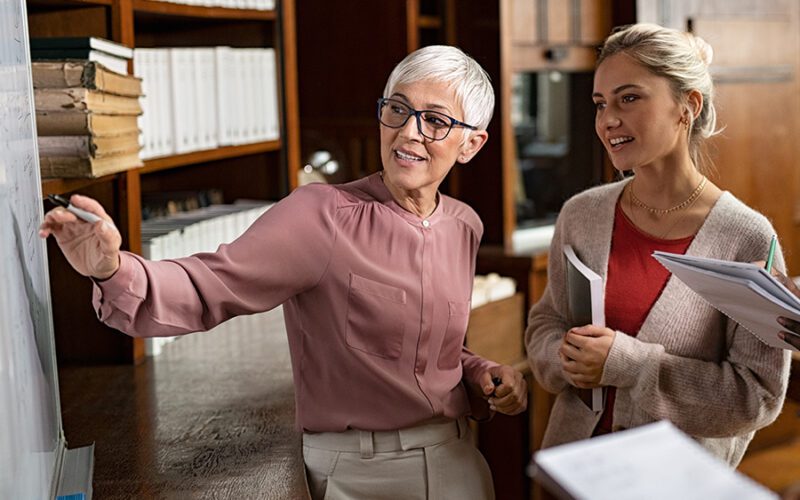 Two women in an office, one with gray hair pointing at a whiteboard, the other holding papers and smiling, surrounded by shelves of books.