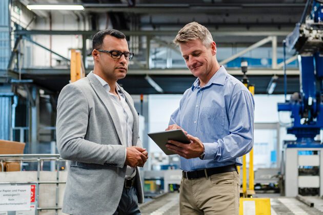 Two men in a factory setting discuss something on a tablet. One wears a gray blazer; the other is in a blue shirt. Industrial equipment is visible in the background.