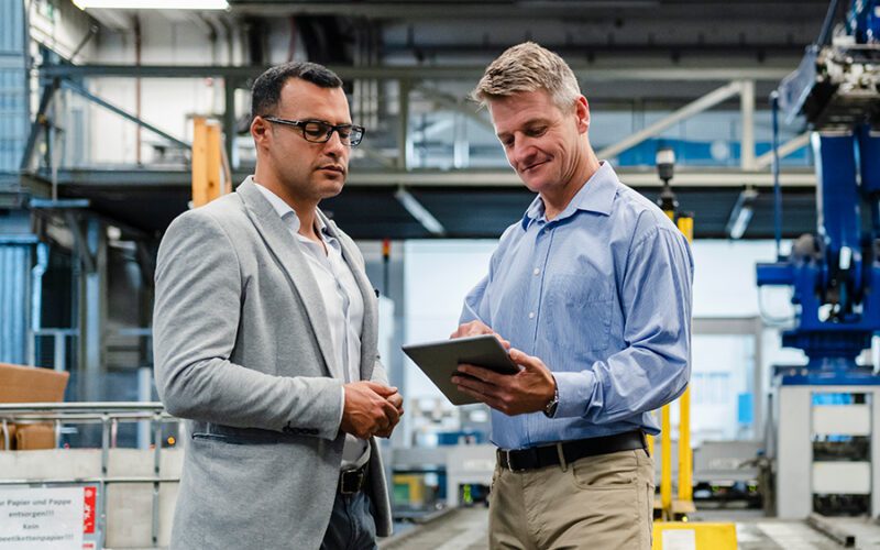 Two men in a factory setting discuss something on a tablet. One wears a gray blazer; the other is in a blue shirt. Industrial equipment is visible in the background.