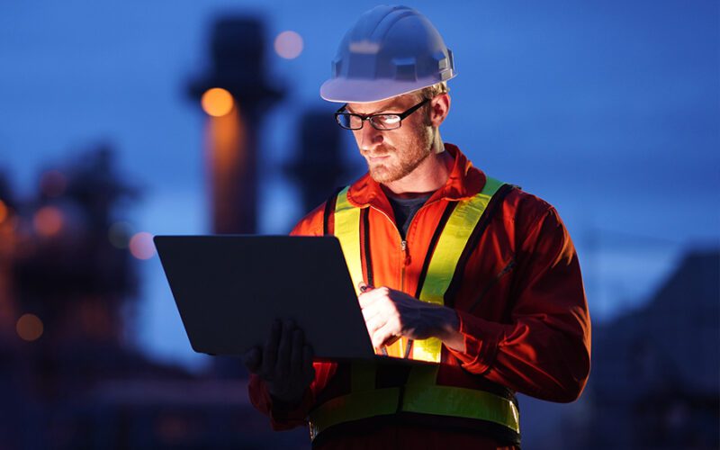 A construction worker in a hard hat and safety vest looks at a laptop outdoors at a construction site in low light conditions.