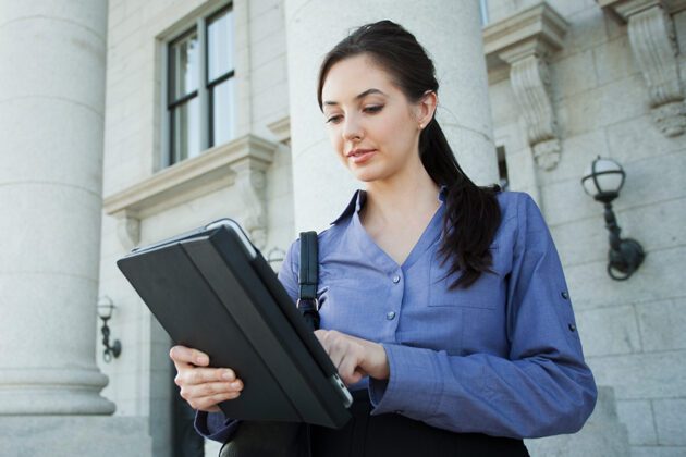 A woman in a blue shirt stands outside a building with columns, using a tablet.