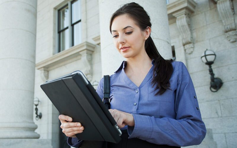 A woman in a blue shirt stands outside a building with columns, using a tablet.