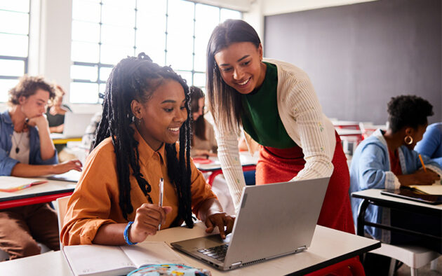 A teacher assists a student working on a laptop in a classroom, with other students seated at desks in the background.