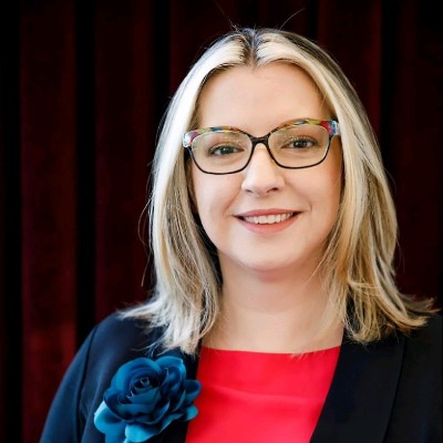 Person with glasses and long blonde hair, wearing a black blazer with a blue flower pin over a red top, stands in front of a dark red curtain.