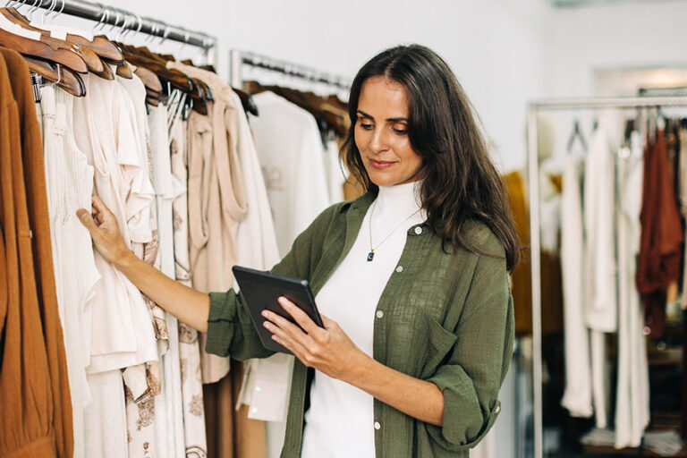 A woman stands by clothing racks, holding a tablet and looking at the screen in a clothing store.