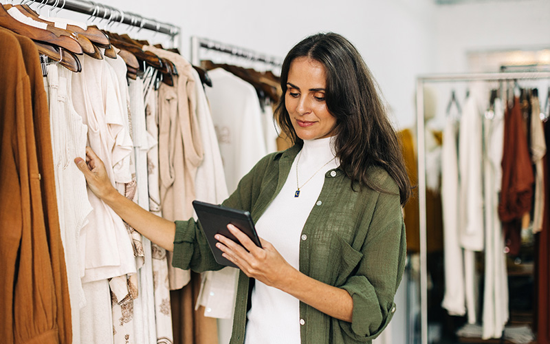 A woman in a green shirt holds a tablet and examines clothes on a rack in a clothing store.