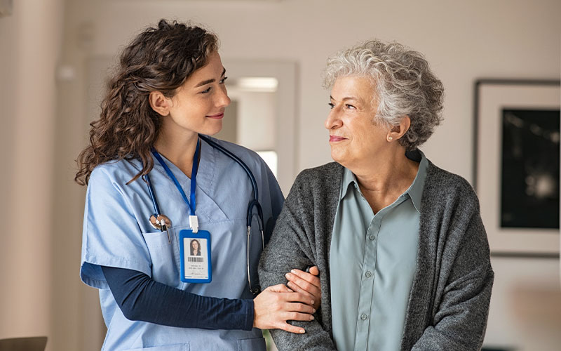 A nurse in blue scrubs with a badge assists an elderly woman in a cardigan. They are standing indoors, smiling at each other.